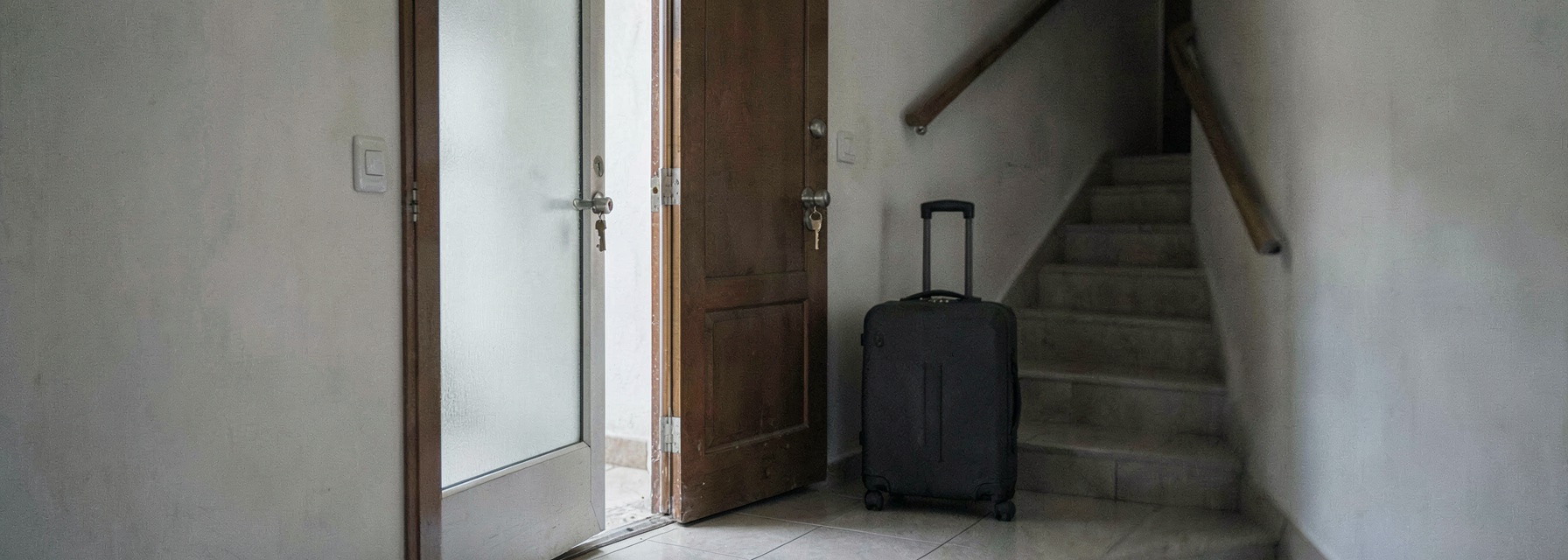 An open apartment door with a key left in the lock and a packed suitcase on the landing